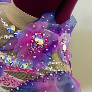 May include: A close-up of a gymnastics leotard with a sheer nude mesh neckline and bodice. The leotard is adorned with iridescent rhinestones and fabric flower appliques in shades of purple, pink, and blue.