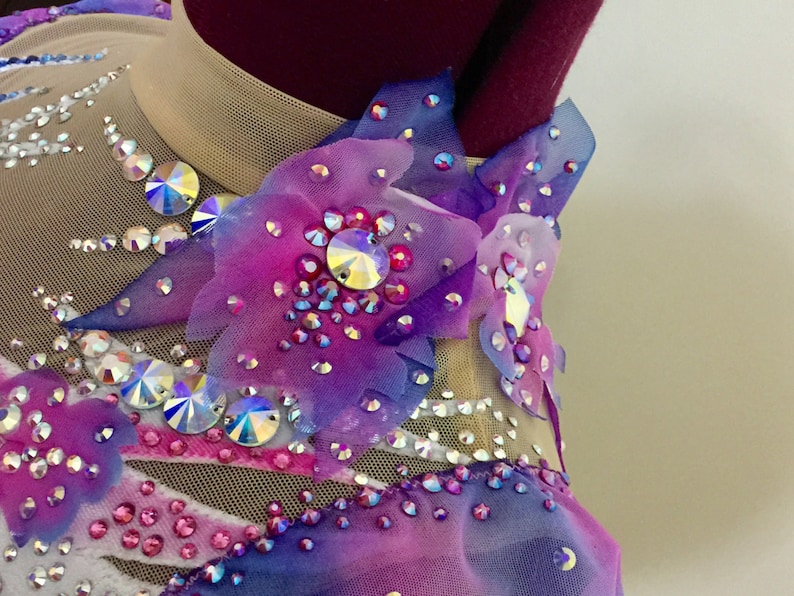 May include: A close-up of a gymnastics leotard with a sheer nude mesh neckline and bodice. The leotard is adorned with iridescent rhinestones and fabric flower appliques in shades of purple, pink, and blue.