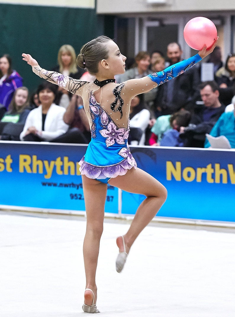 May include: A young gymnast in a blue and purple leotard with floral accents and a pink ball. The leotard has sheer sleeves and is adorned with rhinestones. The gymnast is mid-routine, with arms outstretched and one leg extended.
