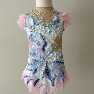 May include: A white and blue leotard with a layered skirt and sheer sleeves. The leotard is embellished with rhinestones and features a design of blue and purple watercolor-style patterns. Pink tulle accents the sleeves and skirt.