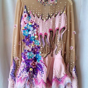 May include: A long-sleeved, light pink leotard with a nude mesh overlay. The leotard is heavily embellished with rhinestones, pearls, and floral appliques in shades of pink, purple, and blue. The design features a high neckline and a skirted bottom.