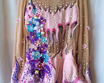 May include: A long-sleeved, light pink leotard with a nude mesh overlay. The leotard is heavily embellished with rhinestones, pearls, and floral appliques in shades of pink, purple, and blue. The design features a high neckline and a skirted bottom.