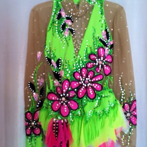 May include: A neon green and pink leotard with floral embellishments. The leotard features a high neckline, long sheer sleeves, and is adorned with large pink flowers, rhinestones, and black accents. The skirt has layers of green, yellow, and pink fabric.