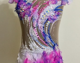 Puede incluir: Un maillot beige con cuello alto y mangas transparentes, adornado con un diseño vibrante de acentos azules, rosas y morados. El disfraz está adornado con pedrería brillante y tela en capas, creando un aspecto dinámico y elegante.