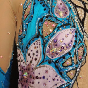 May include: Close-up of a turquoise and beige leotard with floral and abstract designs. The leotard features a large flower with purple and pink petals, and is embellished with rhinestones and sequins. The sleeves are sheer.