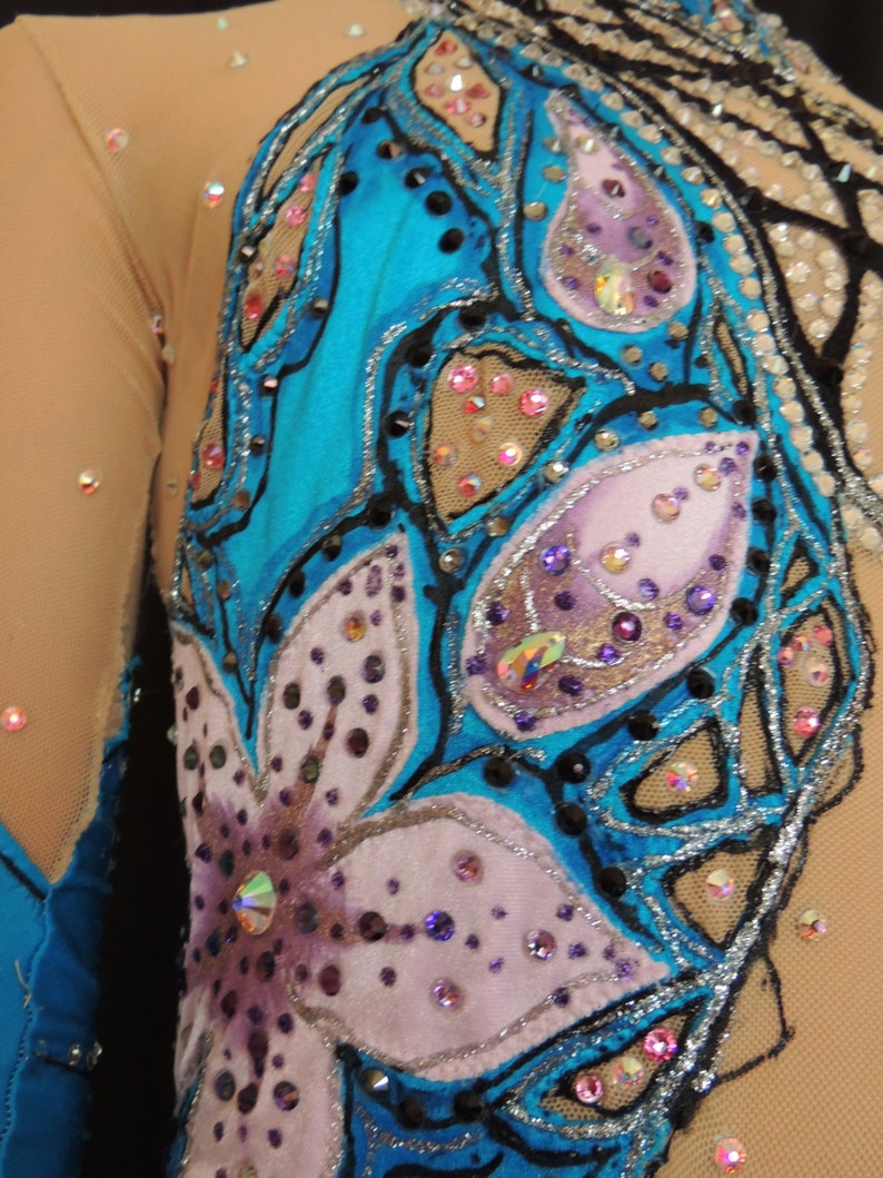 May include: Close-up of a turquoise and beige leotard with floral and abstract designs. The leotard features a large flower with purple and pink petals, and is embellished with rhinestones and sequins. The sleeves are sheer.