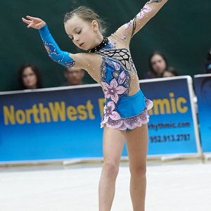 May include: A young gymnast in a blue and pink leotard performs with a hoop. The leotard features floral designs and a sheer overlay. The gymnast is in a graceful pose, with arms extended, during a rhythmic gymnastics competition.