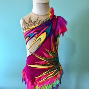May include: A vibrant, one-shoulder leotard with a colorful design. The bodice features a gold crown detail and a mesh neckline. The leotard is adorned with layers of pink, yellow, green, and purple fabric, creating a dynamic and eye-catching look. Perfect for gymnastics or dance.