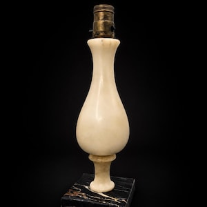 May include: A table lamp base featuring a smooth, cream-colored alabaster body with a curved, vase-like silhouette. The lamp sits on a square, polished black marble base with gold-toned veining and includes a metallic brass-colored socket.