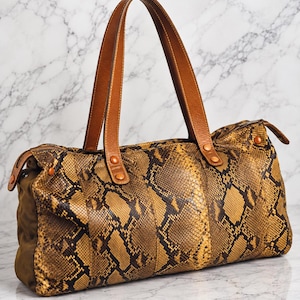 May include: A duffel bag featuring a brown and black snakeskin pattern with tan leather handles. The bag has a top zipper closure, side panels in a solid olive green fabric, and copper-toned metal rivets securing the handles to the main body.