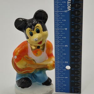 May include: A vintage Mickey Mouse figurine, painted in bright colors, holding a guitar. The figure is approximately 5 inches tall. The figurine is wearing an orange jacket, blue pants, and a white shirt.