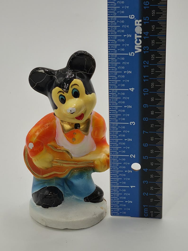 May include: A vintage Mickey Mouse figurine, painted in bright colors, holding a guitar. The figure is approximately 5 inches tall. The figurine is wearing an orange jacket, blue pants, and a white shirt.