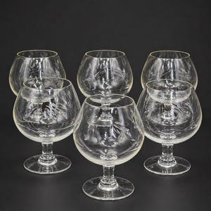 May include: Six clear brandy snifters with etched floral designs. The glasses have a rounded bowl shape, a short stem, and a wide base. The background is a solid black.