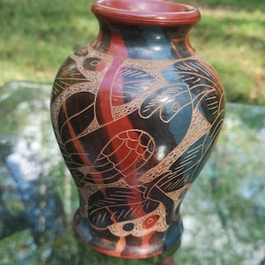 May include: A handcrafted ceramic vase with a unique design. The vase features a dark red rim and base, with a black and red body adorned with etched patterns. The patterns include stylized bird and leaf motifs, creating a decorative and artistic piece.
