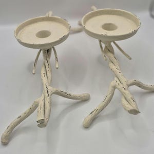 May include: Two off-white candle holders with a distressed finish. The holders have a round top with a central hole for a candle and are supported by twisted, branch-like legs. The candle holders are a decorative home accent.