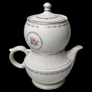 May include: White porcelain tea set with a round top and a lower, wider base. The set features gold trim and a floral design with pink roses and blue accents. The lid has a decorative finial.