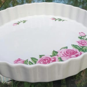 May include: White ceramic quiche dish with a scalloped edge and a floral rose design. The dish features pink roses and green leaves, adding a touch of elegance to any kitchen. Perfect for baking and serving.