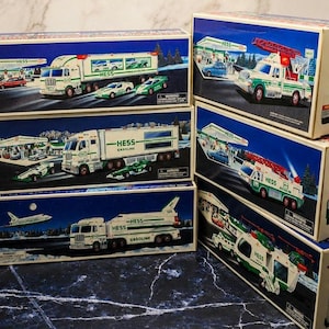 May include: A collection of six rectangular cardboard boxes for Hess toy trucks, each featuring detailed illustrations of white and green tanker trucks, emergency vehicles, and race cars set against snowy, nighttime gas station backgrounds.