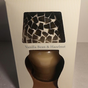 May include: A boxed candle from Shaker Hill Candles. The box is cream-colored with vertical stripes. The front of the box has a window showing a candle with a mosaic-style top and a brown glass jar. The text "Vanilla Bean & Hazelnut" is visible.