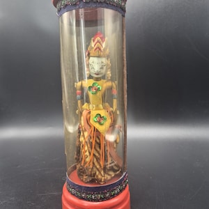 May include: A vintage Indonesian Wayang Golek puppet in a clear cylindrical display case. The puppet has a painted face, a yellow and orange costume, and is mounted on a red base. The case has a red top and is trimmed with decorative fabric.