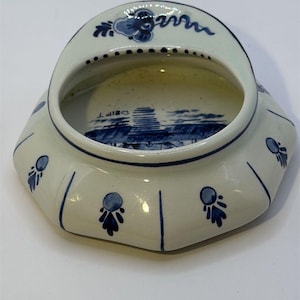 May include: A white ceramic dish with blue floral and landscape designs. The dish has a unique shape with a curved top and a flat base. The rim is decorated with blue floral patterns, and the interior features a blue landscape scene.