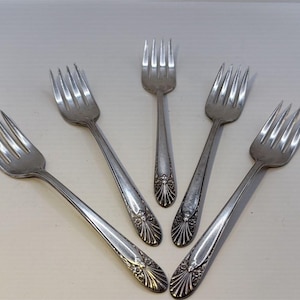 May include: Five silver forks with a decorative floral design on the handles. The forks are arranged in a fan shape, showcasing their tines and the detailed pattern. The silverware is likely for a formal dining setting.