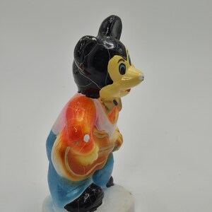 May include: A vintage figurine of a cartoon mouse character. The mouse has a black head, yellow face, and is wearing a colorful outfit of orange, pink, and blue. The figurine is standing on a white base.
