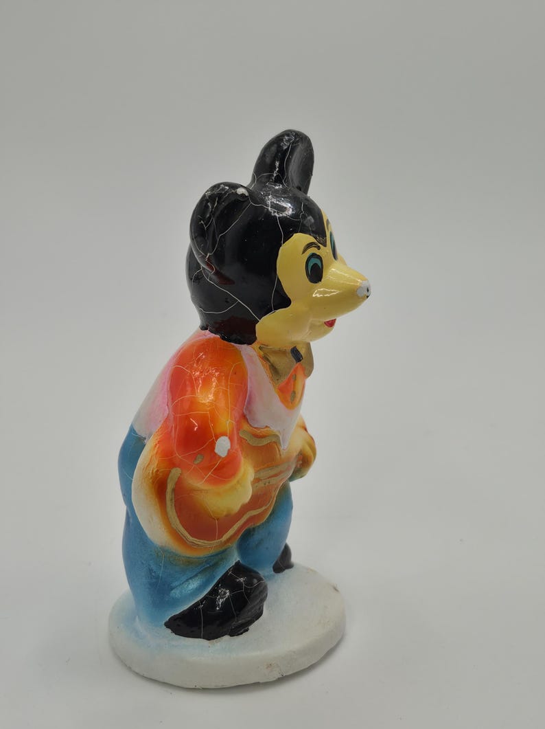 May include: A vintage figurine of a cartoon mouse character. The mouse has a black head, yellow face, and is wearing a colorful outfit of orange, pink, and blue. The figurine is standing on a white base.