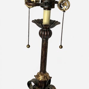 May include: A table lamp with a dark brown and gold finish, featuring a central column with vertical fluting, a decorative base with leaf-shaped accents, two light sockets with pull chains, and a pointed finial at the very top of the stem.