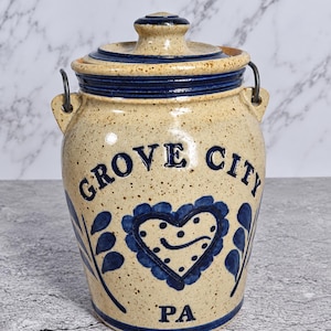 May include: A speckled beige stoneware crock with a dark blue lid and metal wire handle. The front features the text GROVE CITY PA in dark blue lettering, centered above a dark blue heart design with a dotted pattern and leaf accents.