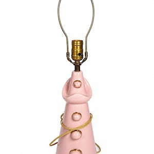May include: A pink ceramic table lamp with a brass neck and a gold-colored wire. The lamp has a unique design with three decorative gold-colored circles and a bulb socket. The lamp is approximately 15 inches tall.