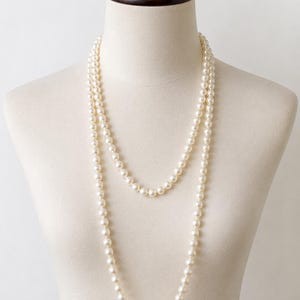 May include: A long, double-strand necklace featuring uniform, round, cream-colored pearls. The necklace is draped in two loops, with the shorter strand resting higher on the chest and the longer strand hanging lower, creating a layered appearance.