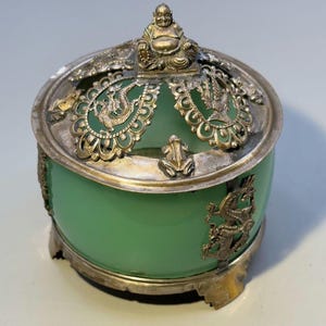 May include: A round green glass container with a silver-toned metal lid and base. The lid features an ornate openwork design with a small seated figure at the top, surrounded by dragon motifs and small frog figures attached to the metal rim.