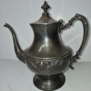 May include: A silver-colored teapot with a curved spout and ornate handle. The teapot has a decorative floral pattern around the base and a lid with a small finial. The teapot sits on a round base.