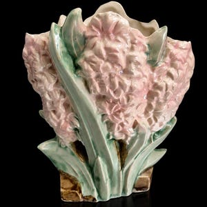 May include: A ceramic vase shaped like a cluster of pink hyacinth flowers with green leaves. The vase has a textured surface and sits on a brown base. The background is black.
