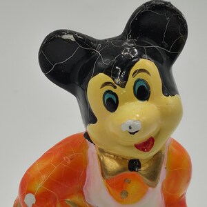 May include: A vintage Mickey Mouse figurine with a yellow face, black ears, and an orange and white outfit. The figurine has a gold bow tie and a red mouth. The paint shows signs of age and wear.