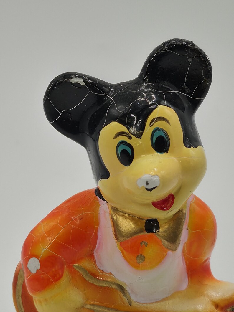 May include: A vintage Mickey Mouse figurine with a yellow face, black ears, and an orange and white outfit. The figurine has a gold bow tie and a red mouth. The paint shows signs of age and wear.