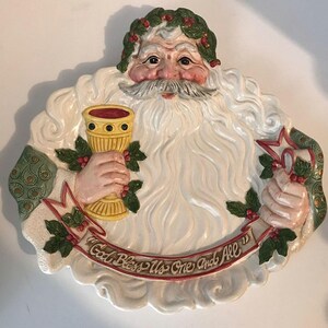 May include: A ceramic Santa Claus figure with a white beard and a green holly wreath. He holds a golden chalice and a star-shaped ornament. The banner reads "God Bless Us One and All."