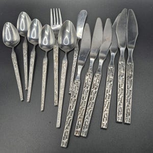 May include: A collection of vintage silverware, including spoons, forks, and knives. The stainless steel utensils have a textured, geometric pattern on the handles. The set includes various sizes of spoons, forks, and knives, perfect for a formal dining setting.