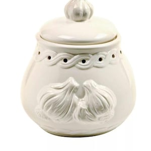 May include: A white ceramic garlic keeper with a lid and a garlic-shaped handle. The jar features a decorative wavy pattern and embossed garlic cloves. The keeper is designed to store garlic and keep it fresh.