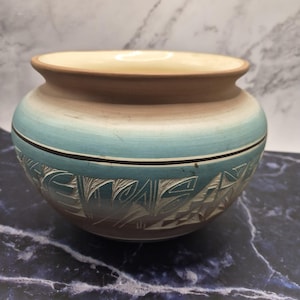 May include: A ceramic pot with a wide, rounded body and a flared rim. The upper portion is a smooth, light beige, transitioning into a vibrant turquoise band. The lower section features intricate, carved geometric patterns in a dark brown clay.