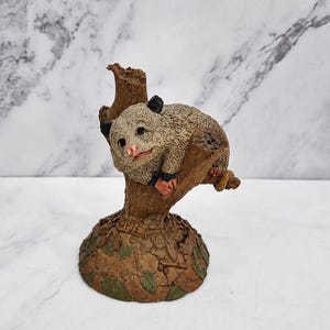 May include: A decorative figurine of a grey and white opossum resting on a brown tree stump. The base is textured with sculpted leaves and features the name Opie embossed on the front. The opossum has a pink nose, black ears, and paws.