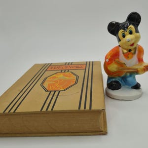 May include: A vintage book with the title "The Fairy" and a decorative illustration, next to a small figurine of a cartoon mouse playing a guitar. The figurine is wearing an orange vest and blue pants.