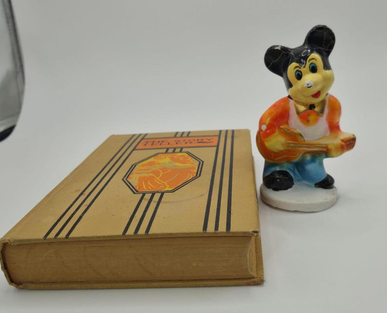 May include: A vintage book with the title "The Fairy" and a decorative illustration, next to a small figurine of a cartoon mouse playing a guitar. The figurine is wearing an orange vest and blue pants.