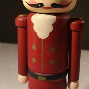May include: A wooden nutcracker figurine in the shape of a soldier. The figurine has a blue hat, red jacket with gold accents, and a black belt. The face is painted with a mustache and rosy cheeks. The base is black.
