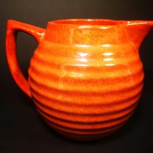 May include: A vibrant orange ceramic pitcher with a ribbed design. The pitcher has a handle and a spout for pouring. The glossy finish reflects light, highlighting the texture. This decorative pitcher is suitable for serving beverages.