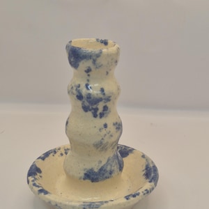 May include: A handmade ceramic candle holder with a cream-colored base and blue speckled design. The candle holder has a unique, curvy shape and sits on a matching saucer. The overall design is rustic and artistic.