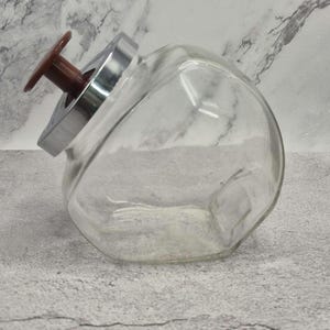 May include: A clear glass storage jar with a slanted opening, featuring a metallic silver lid and a brown plastic knob handle. The container has a wide, rounded body and a flat base, designed to sit at an angle for easy access to contents.