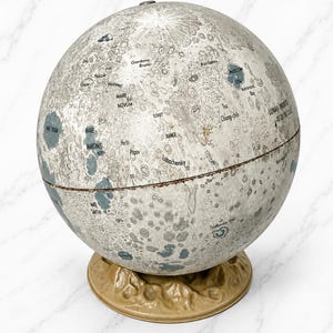 May include: A spherical moon globe featuring a detailed gray and white lunar surface map with labeled craters and seas, including text such as Mare Novum, Mare Marginis, and Soviet Range. The globe rests on a textured, tan-colored circular base.