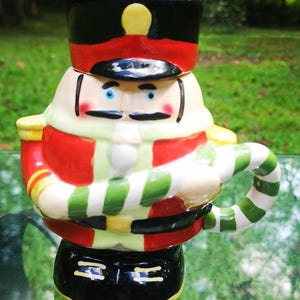 May include: A ceramic mug shaped like a nutcracker soldier. The mug features a black hat with a yellow accent, a red jacket, and a green and white striped handle. The mug is a festive holiday decoration.